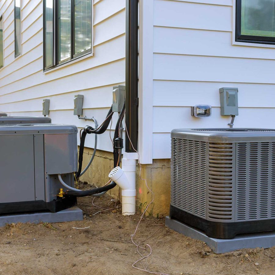 Four air conditioning units sit beside a white house, connected with pipes and electrical lines against a gravelly ground and windows.